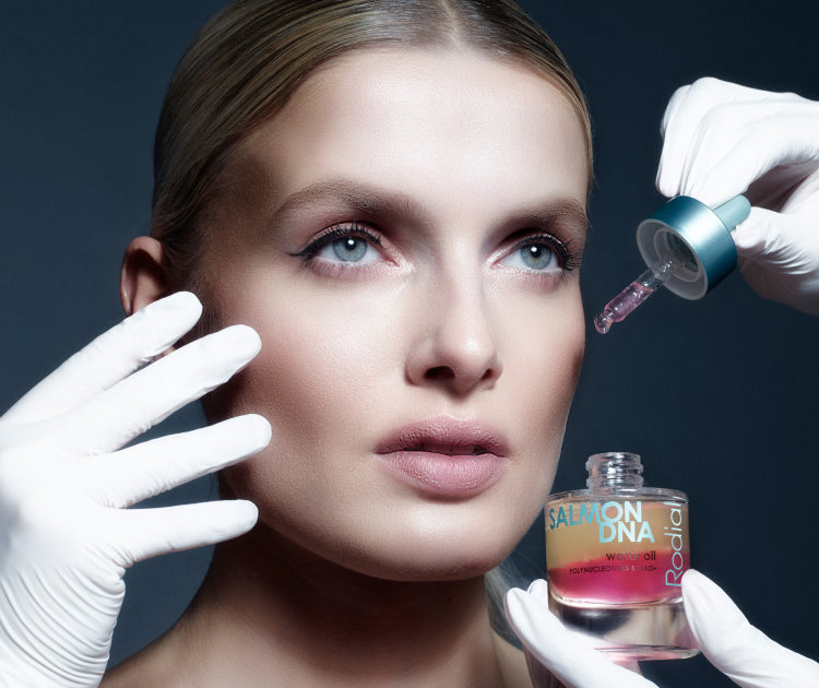 Woman with gloves applying a dropper of serum to her face, with a bottle of serum in the foreground.